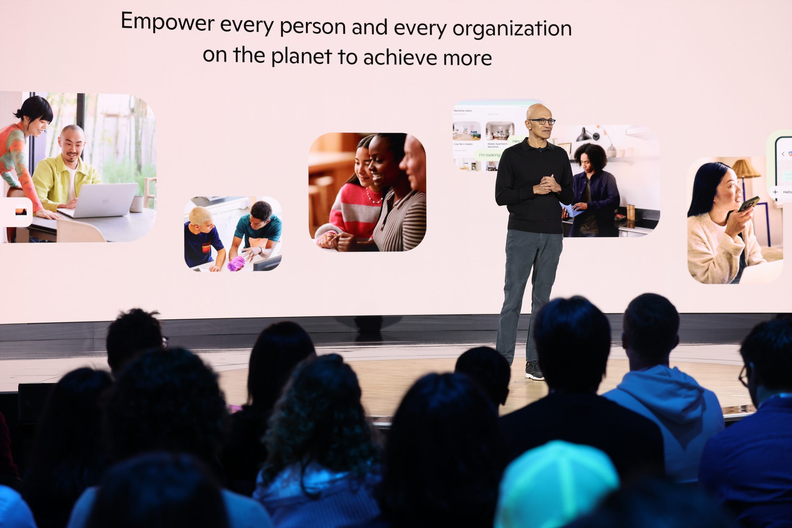 A man stands on a stage in front of a screen with photos and text that reads, “Empower every person and every organization to achieve more” while an audience looks on.